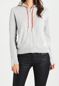 Szara bluza rozpinana na zamek z pomarańczowymi sznurkami, ściągaczami na mankietach i na dole. Gładka faktura i casualowy design, w połączeniu z czarnymi, dopasowanymi spodniami.