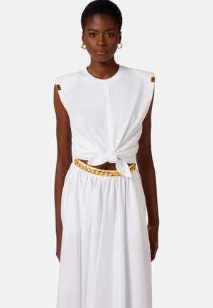 Donna che indossa un top bianco senza maniche legato in vita con cintura a catena dorata e gonna abbinata, in piedi su sfondo semplice.