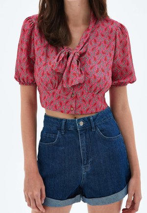 Kobieta nosząca ciemne jeansowe szorty z wysokim stanem, podwinięte, oraz różowy krótki top z krótkim rękawem, z wzorem paisley i wiązaną kokardą z przodu.