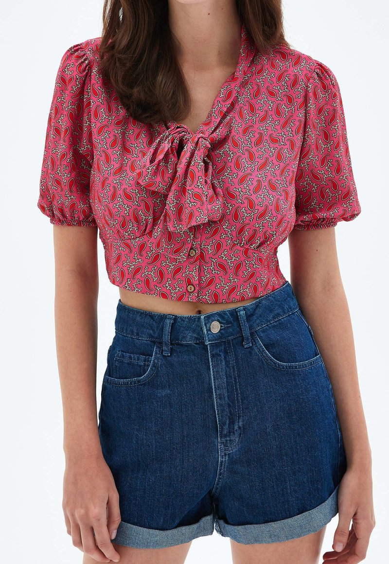 Femme portant un short en denim foncé taille haute roulé et un crop top rose à manches courtes avec un motif cachemire et un nœud à l'avant.