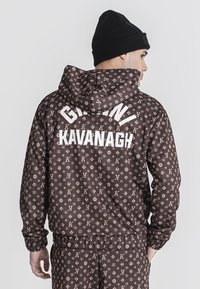 Chaqueta marrón con un patrón de logo repetido en un color beige claro, con capucha y puños elásticos. La parte trasera muestra "GIANNI KAVANAGH" en texto blanco.