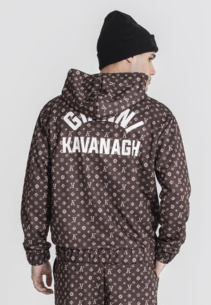 Veste marron avec un motif de logo répété en beige clair, dotée d'une capuche et de poignets élastiques. Le dos affiche "GIANNI KAVANAGH" en lettres blanches.