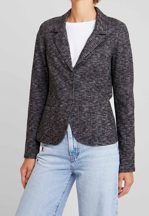 Kobieta ubrana w dopasowaną ciemnoszarą kratowaną marynarkę na dwa guziki, w połączeniu z jasnoniebieskimi dżinsami i białym topem.