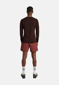 Mann steht mit dem Rücken zur Kamera, trägt ein braunes Langarmshirt, bordeauxrote Shorts, weiße Socken mit "OFF DUTY GEAR" und beige Sneakers.