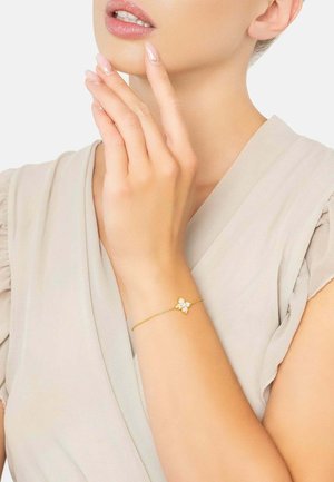 Goldene Kettenarmband mit einem floralen Design und drei weißen Steinen in der Mitte; glatte Textur, filigranes Design, am Handgelenk getragen.