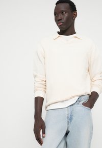 Lys beige tekstureret sweater med V-hals og lange ærmer, båret over en hvid T-shirt, parret med lyseblå denimjeans.