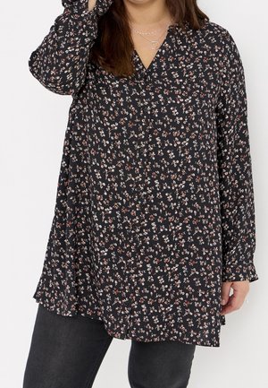 Mujer con blusa negra de manga larga con pequeño estampado floral blanco y rojo y pantalones gris oscuro, de pie frente a un fondo blanco.
