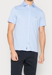 Camisa de manga corta azul claro con cuello abotonado, hecha de tela suave. Cuenta con un pequeño emblema de logo y una etiqueta en el dobladillo con los colores de la marca.