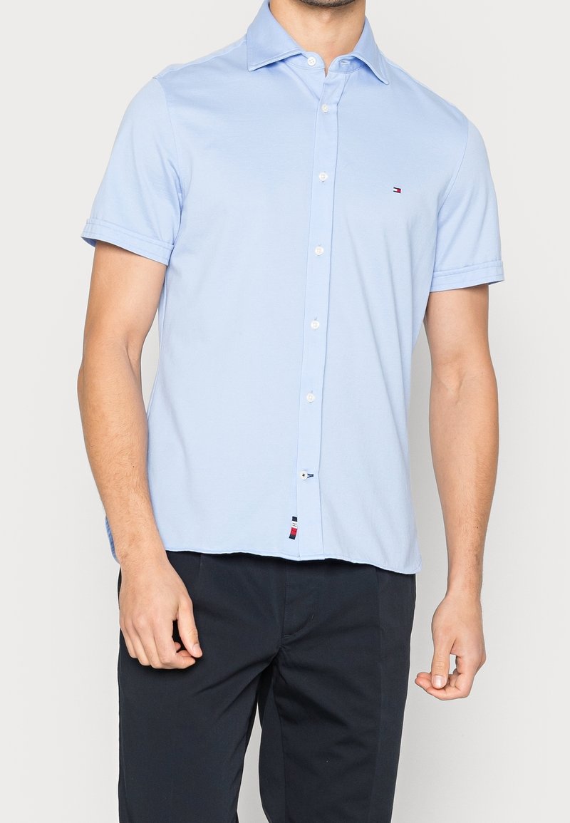 Camisa de manga corta azul claro con cuello abotonado, hecha de tela suave. Cuenta con un pequeño emblema de logo y una etiqueta en el dobladillo con los colores de la marca.