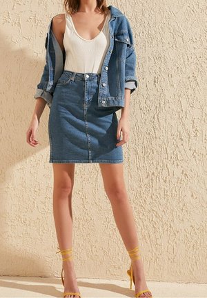 Femme portant un débardeur blanc, une veste en jean tombant des épaules, une jupe en jean et des sandales jaunes à talons hauts, debout contre un mur beige texturé.