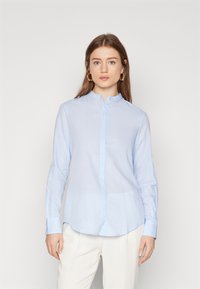 Camicia azzurro chiaro con colletto alla mandarino, maniche lunghe e orlo curvo. Realizzata in un tessuto leggero con una texture liscia.