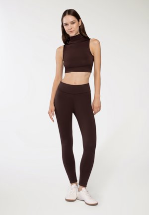 Top corto marrone con collo alto e leggings marroni abbinati, entrambi realizzati in materiale elasticizzato. Indossato con scarpe da ginnastica bianche.