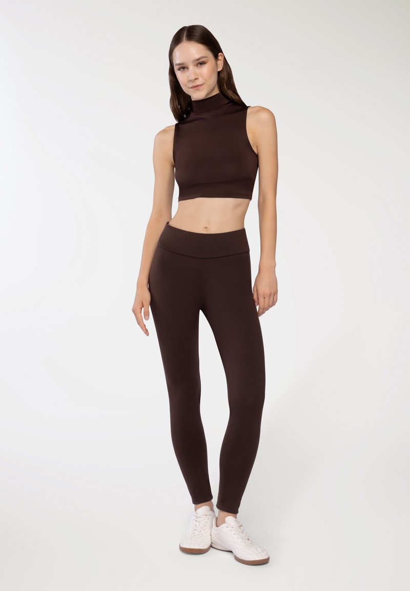 Top corto marrone con collo alto e leggings marroni abbinati, entrambi realizzati in materiale elasticizzato. Indossato con scarpe da ginnastica bianche.