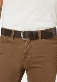 Brauner Leder-Gürtel mit einer silbernen Metallschnalle, der eine glatte Textur aufweist und mehrere Löcher zur Anpassung hat, getragen über braunen Hosen.