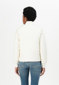 Weiße gesteppte Bomberjacke mit hohem Kragen und gerippten Bündchen, kombiniert mit blauen schmal geschnittenen Jeans. Der Stoff hat eine weiche Textur und ein Diamantmuster.