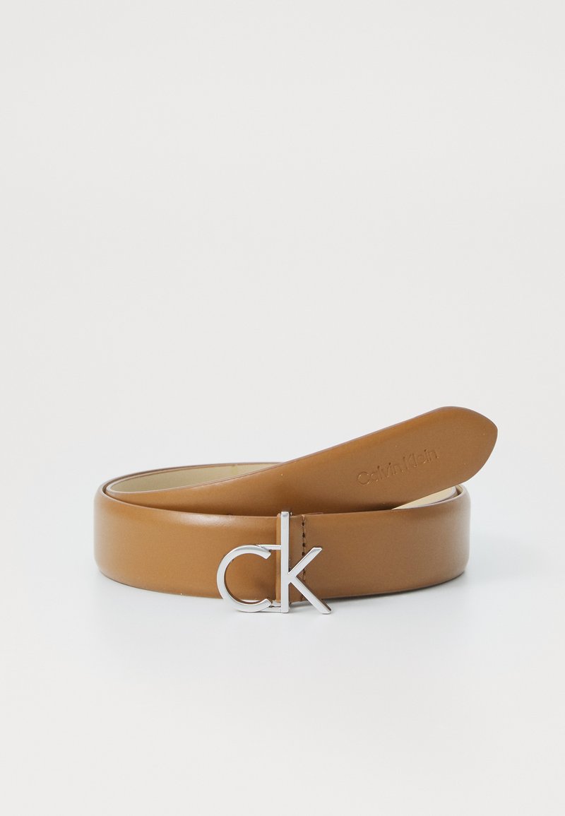 Ceinture en cuir tan avec une boucle en argent poli en forme de "CK" et une texture lisse. Le design est simple et épuré avec des accents minimaux.