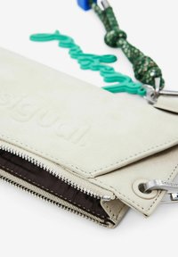 Crèmekleurige portemonnee van suède, met een zilveren ritssluiting, ingeslagen logo en een kleurrijk sleutelhanger bevestigd, met textuuraccenten.