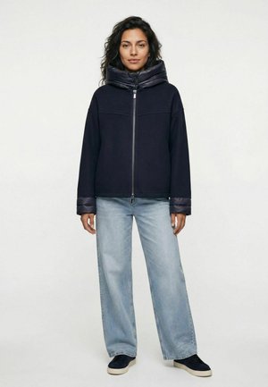 Frau mit dunkel lockigem Haar steht und trägt eine dunkle Jacke mit Reißverschluss, gepolstertem Kragen und Bündchen, hellblaue Jeans mit weitem Bein und schwarze Sneakers.