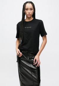 Juoda medvilninė trumpomis rankovėmis marškinėliai su prekės ženklo "PINKO" pavadinimu kontrastingu atspaudu. Derinami su blizgia juoda odine sijonu.
