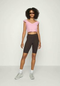 Top cropped rosa com padrão de ilhós e mangas franzidas, combinado com calções de bicicleta castanhos de cintura alta e sapatilhas cinzentas. A modelo usa óculos de sol escuros.