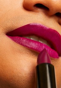 Mørk magenta leppestift på fyldige lepper, med jevn tekstur og definerte kanter. Leppestiftmynte er spiss og synlig med et svart etui.