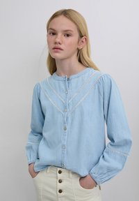 Camisa azul claro de botones, confeccionada en tela suave, con encaje en los bordes, mangas largas abullonadas con puños elásticos y escote redondeado.