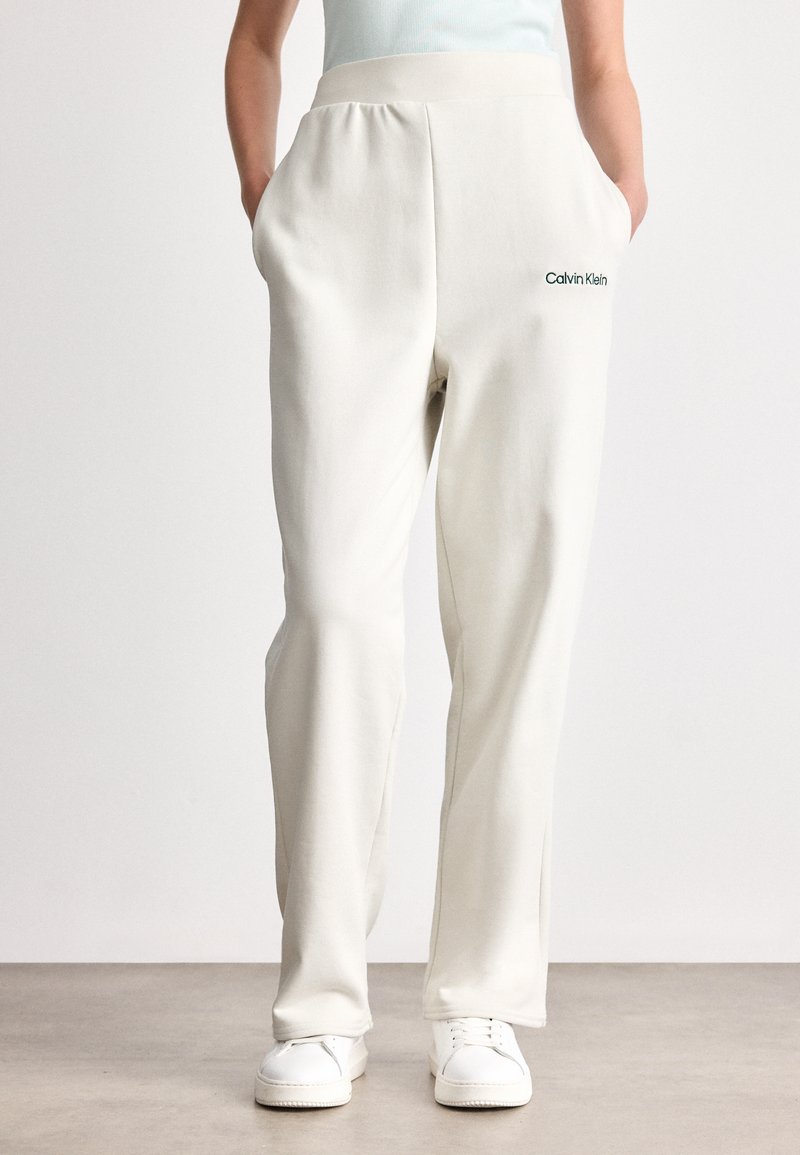 Άτομο που φοράει off-white φόρμες Calvin Klein και λευκά sneakers, στέκεται με τα χέρια στις τσέπες του μπροστά σε απλό φόντο.