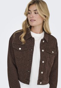 Chaqueta de mezclilla corta con estampado de leopardo marrón y herrajes plateados, con dos bolsillos en el pecho y cierres de botón, llevada sobre una camisa blanca.