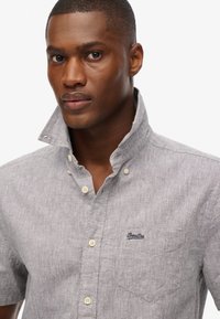 Camicia grigia in chambray a maniche corte con colletto a punta, taschino sul petto e dettaglio del logo. Tessuto texturizzato con bottoni a contrasto.