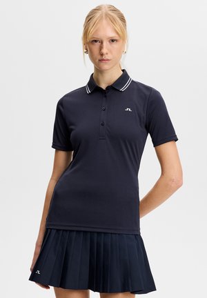 Kobieta o blond włosach, ubrana w granatową koszulkę polo z białymi paskami na kołnierzyku oraz pasującą plisowaną spódnicę, na jednolitym tle.