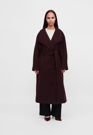 Femme debout portant un long manteau ceinturé marron foncé sur un pantalon noir et des chaussures marron à talons aiguilles pointues, sur un fond clair uni.