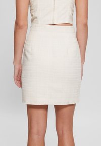 Jupe courte et ajustée en tissu texturé beige clair avec un éclat subtil, dotée d'une fermeture éclair dissimulée à l'arrière et sans embellissements visibles.