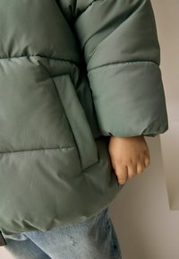 Green puffer jacket with a matte finish, featuring a large pocket on the side and round, thick sleeves. Worn over light blue distressed jeans.