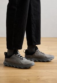 Baskets de sport grises avec une tige en cuir texturé, des accents noirs et une semelle en caoutchouc épaisse, portées avec un pantalon sombre et des chaussettes grises claires.