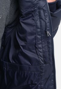 Marineblå dunjakke med blank, glat overflade, der har en lynlås lomme og elastiske detaljer. Struktureret grå striktrøje underneden synlig.