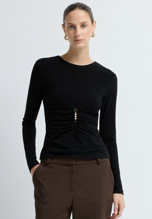 Sort langærmet top med rund halsudskæring og drapering i taljen, prydet med guldfarvet hardware og en dekorativ detalje i midten.