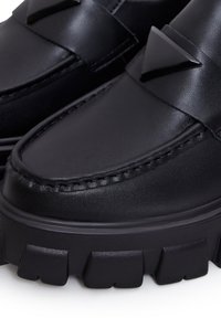 Mocassins en cuir noir épais avec des semelles lug épaisses et un détail en métal triangulaire sur le dessus.