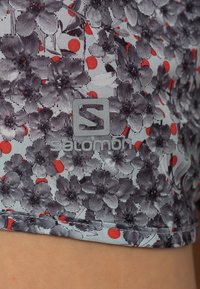 Närbild av grå och svart blommönstrad tyg med röda prickar, med Salomon-logotypen tryckt på materialet.