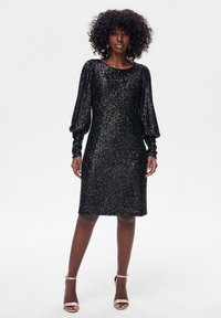 Kobieta o kręconych włosach, ubrana w czarną, cekinową sukienkę do kolan z bufkami na długich rękawach oraz beżowe, odkryte buty na obcasie na białym tle.