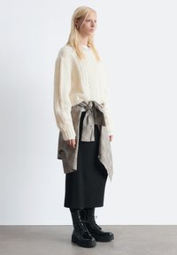 Cremfarbener Kabelstrickpullover mit abgefallenen Schultern, über einem karierten Hemd, das in der Taille gebunden ist, kombiniert mit einem schwarzen Midikleid und klobigen schwarzen Stiefeln.