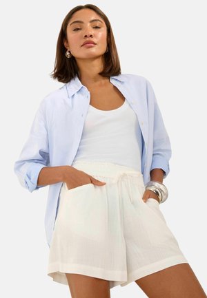 Kvinde iført lyseblå, opknappet skjorte over hvid tanktop og hvide shorts med snørre, med sølv øreringe i hoop-stil og chunky armbånd.