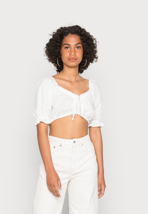Blusa branca curta com mangas bufantes e decote com cordão. Combinada com calças de cintura alta de cor clara. Textura de tecido suave.