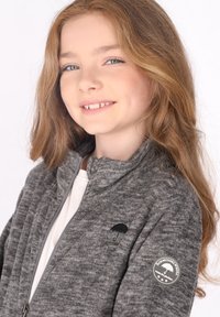 Giacca grigia con zip realizzata in tessuto texturizzato, caratterizzata da un logo a ombrello nero sul petto e un badge rotondo sulla manica sinistra.