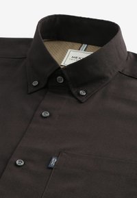 Chemise marron foncé boutonnée avec col boutonné et étiquette visible sur la poche avant sur fond blanc.