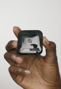 Zwarte, glanzende compacte product dat in een hand wordt vastgehouden, met een vierkante vorm en het logo "KIKO" op de voorkant. Gladde oppervlakte, minimalistisch ontwerp.