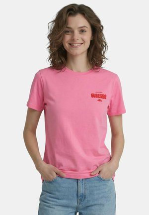 Jeune femme aux cheveux bruns bouclés sourit, portant un t-shirt rose à manches courtes et un jean bleu clair, les mains dans les poches.