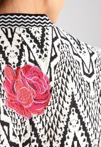 Svartvitt mönstrad tyg med en framträdande röd och rosa broderad blomma nära kragen på en persons axel.