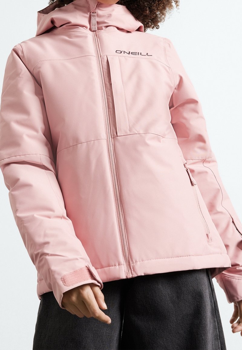 Personne portant une veste zippée rose clair O'Neill avec capuche et sangles velcro aux poignets, associée à un pantalon foncé, sur un fond uni.
