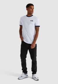 Valge T-särk, millel on tumesinine soonikkoes krae ja varrukad, mustad teksad ning valged sneaksid, millel on tumesinised aktsendid. Mudel seisab käed taskutes.