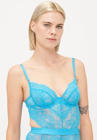 Turquoise lace bralette with underwire cups, intricate floral pattern, adjustable straps, and a smooth waistband detail.